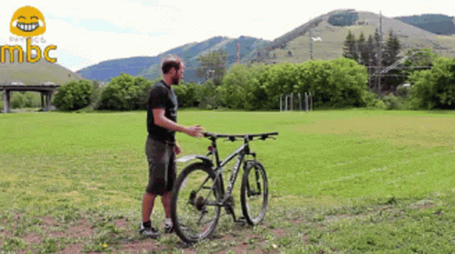 A Man Stands Next To A Bicycle In A Grassy Field With Mountains In The Background And The Word Mbc In The Corner GIF
