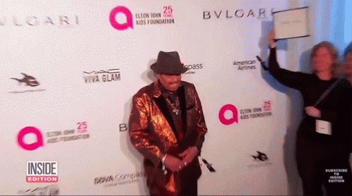 A Man Stands On A Red Carpet In Front Of A Wall That Says Bvlgari GIF