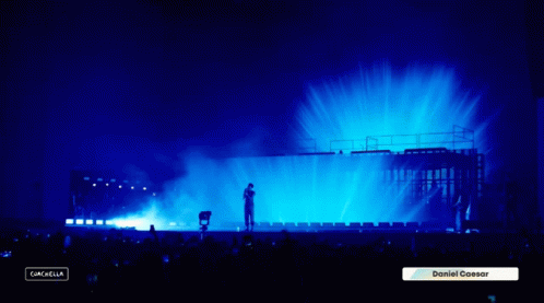 A Man Stands On A Stage With A Blue Light Behind Him And A Sign That Says Daniel Caesar GIF
