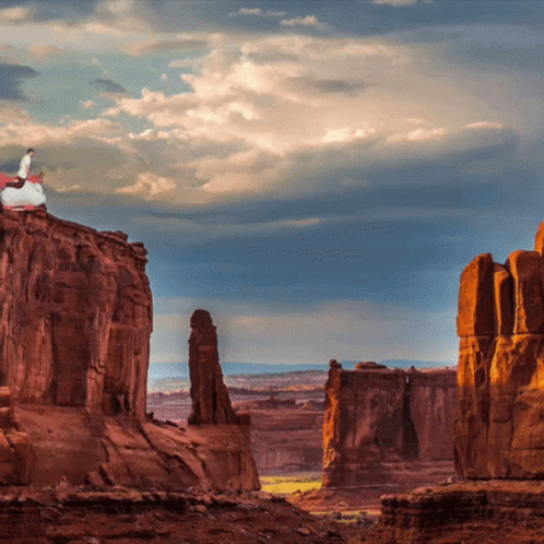 A Man Stands On Top Of A Rocky Cliff Overlooking A Canyon GIF