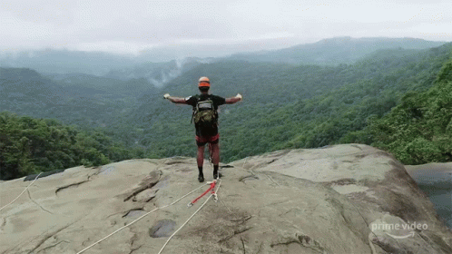 A Man Stands On Top Of A Rocky Cliff With His Arms Outstretched In Front Of A Lush Green Forest .. GIF
