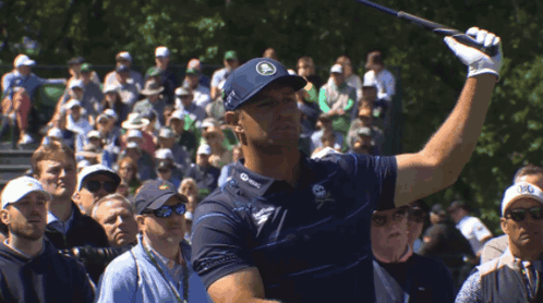 A Man Swings A Golf Club In Front Of A Crowd That Includes A Man Wearing A Hat That Says R On It GIF