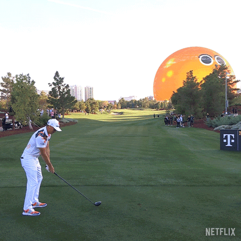 A Man Swings A Golf Club On A Golf Course With An Orange Balloon In The Background GIF