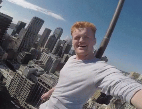 A Man Taking A Selfie On Top Of A Building With A City Skyline In The Background GIF