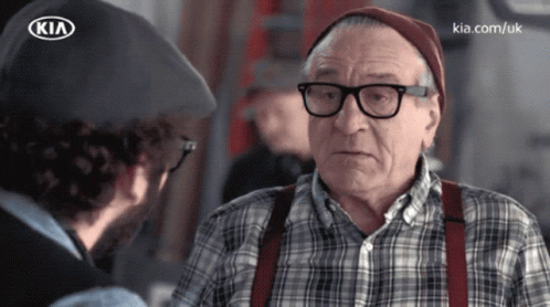 A Man Talking To An Older Man With Glasses And Suspenders And A Kia Logo On His Hat GIF