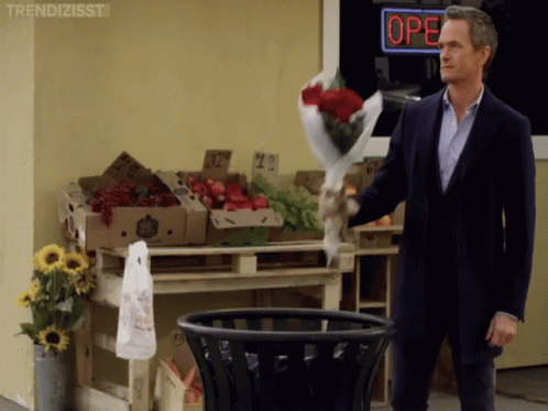 A Man Throws A Bouquet Of Roses Into A Trash Can In Front Of A Store That Is Open GIF