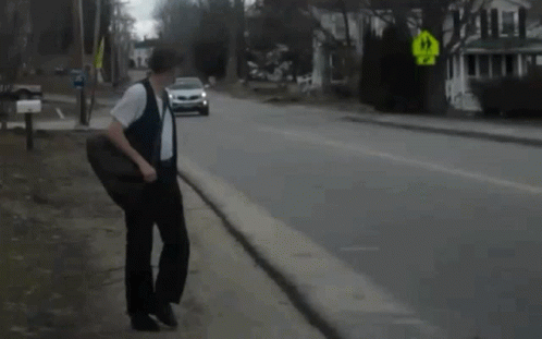 A Man Walking Down A Street With A Car Behind Him And A Yellow Sign That Says Right Turn GIF