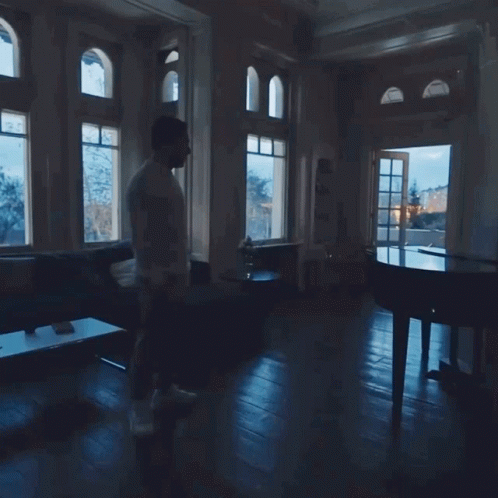 A Man Walking In A Dark Room With A Piano In The Background GIF