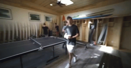 A Man Walking In A Room With A Ping Pong Table In The Background GIF
