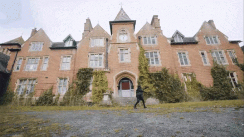 A Man Walking In Front Of A Large Brick Building With A Clock Tower GIF