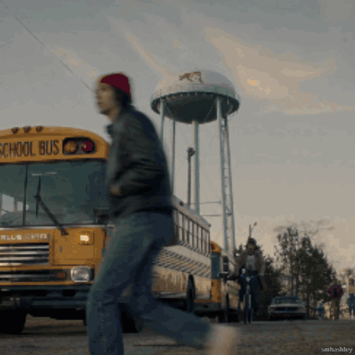A Man Walking In Front Of A School Bus With A Water Tower In The Background GIF