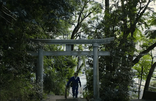 A Man Walking Up A Set Of Stairs Under A Torii Gate With Trees In The Background GIF