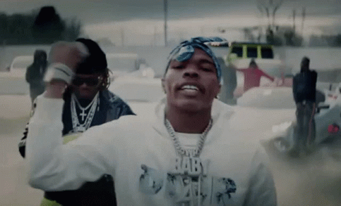 A Man Wearing A Bandana And A Hoodie With The Word Baby On It Is Dancing In A Parking Lot . GIF