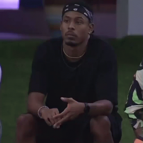 A Man Wearing A Bandana Is Sitting On The Grass With His Hand To His Chin . GIF