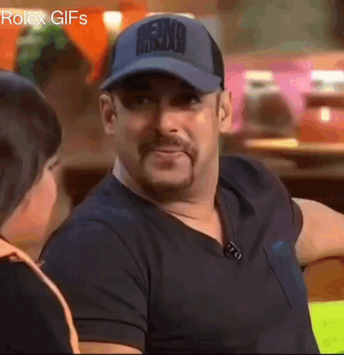 A Man Wearing A Baseball Cap And A Black Shirt Is Smiling While Talking To A Woman . GIF
