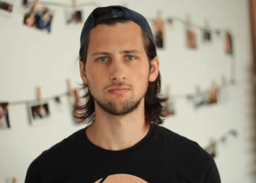 A Man Wearing A Baseball Cap And A Black Shirt Stands In Front Of A Wall With Pictures Hanging On It GIF