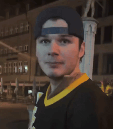 A Man Wearing A Baseball Cap And A Black Shirt With A Yellow Collar Looks At The Camera GIF