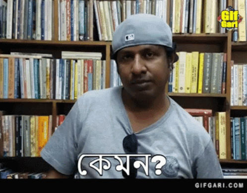 A Man Wearing A Baseball Cap And A Gray Shirt Is Standing In Front Of A Bookshelf And Says Komon GIF