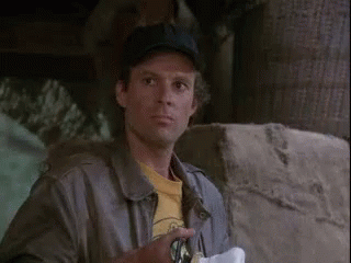 A Man Wearing A Baseball Cap And A Leather Jacket Is Standing Next To A Stone Wall . GIF