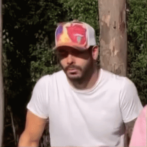 A Man Wearing A Baseball Cap And A White Shirt Is Standing In Front Of A Wooden Pole . GIF