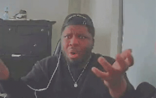 A Man Wearing A Baseball Cap Is Sitting At A Desk With His Hands Folded In Front Of His Face . GIF