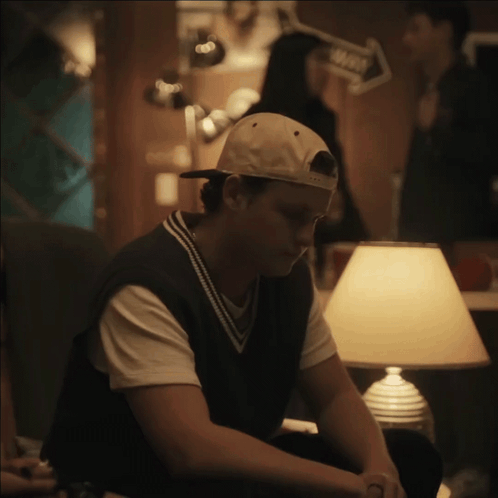 A Man Wearing A Baseball Cap Sits In Front Of A Lamp In A Dark Room GIF