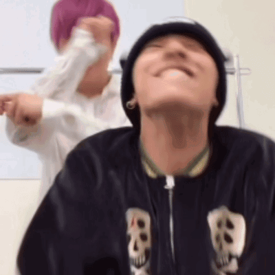 A Man Wearing A Beanie And A Jacket With Skulls On It Is Smiling While Another Man Stands Behind Him . GIF