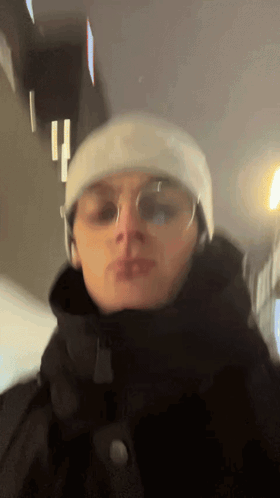 A Man Wearing A Beanie And Glasses Is Standing In A Dark Room GIF