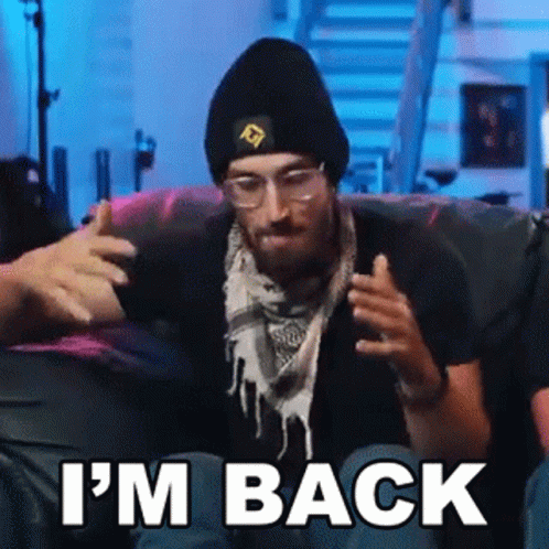 A Man Wearing A Beanie And Scarf Is Sitting On A Couch With The Words `` I 'M Back '' . GIF