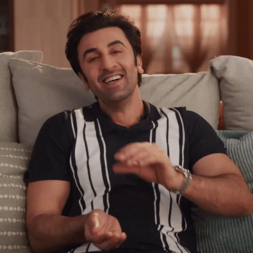 A Man Wearing A Black And White Striped Shirt Is Smiling While Sitting On A Couch GIF