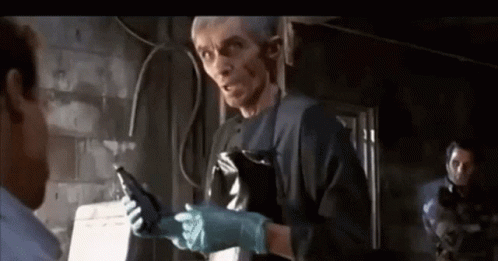 A Man Wearing A Black Apron And Gloves Is Talking To Another Man In A Dark Room . GIF