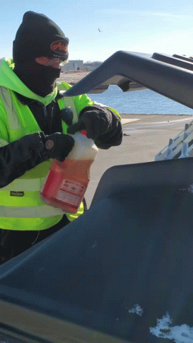A Man Wearing A Black Beanie And A Yellow Vest Holds A Gallon Of Orange Liquid In His Hand GIF