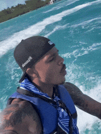 A Man Wearing A Black Hat And A Blue Vest Is Riding A Boat GIF