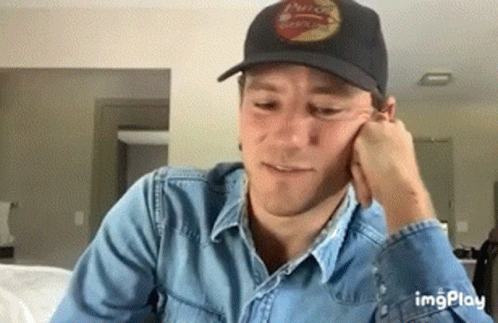 A Man Wearing A Black Hat And A Denim Shirt Is Sitting On A Couch . GIF