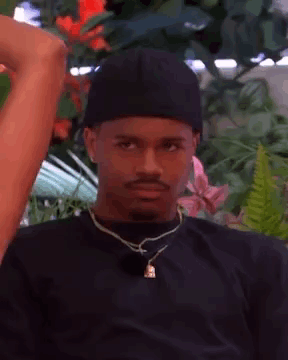 A Man Wearing A Black Hat And A Necklace Is Sitting In Front Of A Flower Garden . GIF