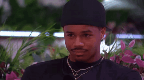 A Man Wearing A Black Hat And A Necklace Is Talking To A Woman In Front Of Flowers . GIF