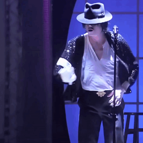 A Man Wearing A Black Hat And A White Shirt Is Standing In Front Of A Microphone GIF