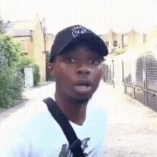 A Man Wearing A Black Hat And A White Shirt Is Standing On A Sidewalk . GIF