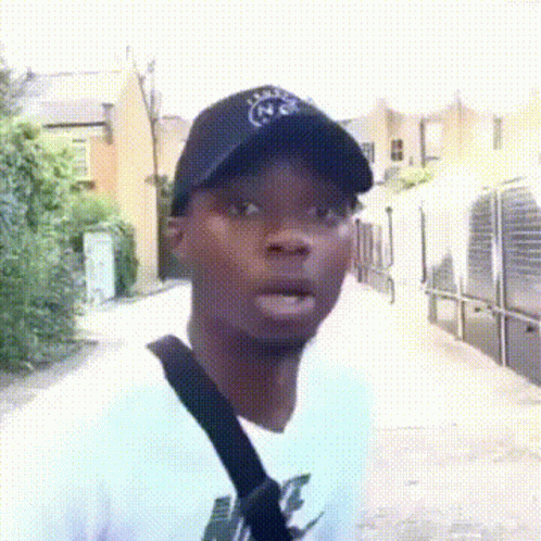 A Man Wearing A Black Hat And A White Shirt Is Standing On A Street . GIF