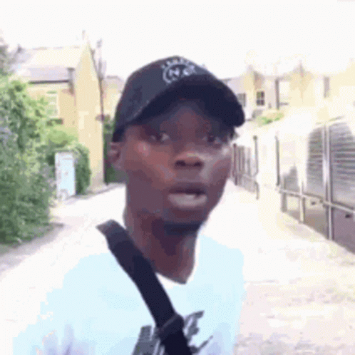 A Man Wearing A Black Hat And A White Shirt Is Standing On A Street . GIF
