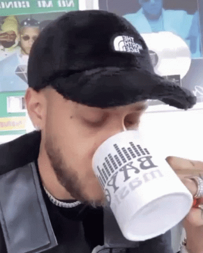 A Man Wearing A Black Hat Is Drinking From A White Mug That Says Bxa On It GIF
