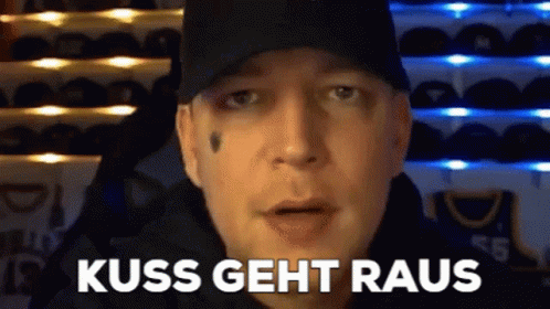 A Man Wearing A Black Hat Is Standing In Front Of A Wall Of Hats And Says Kiss Geht Raus . GIF
