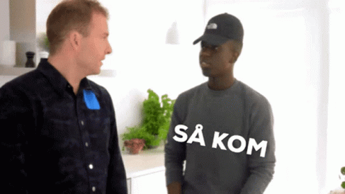 A Man Wearing A Black Hat Talks To Another Man Who Is Wearing A Grey Shirt That Says Sa Kom GIF