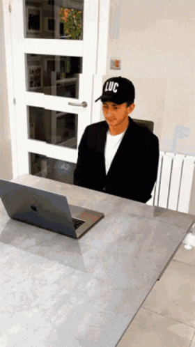 A Man Wearing A Black Hat With Luc On It Sits At A Desk GIF