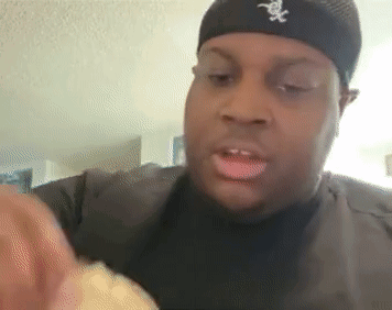 A Man Wearing A Black Hat With The Letter X On It Is Eating A Sandwich . GIF