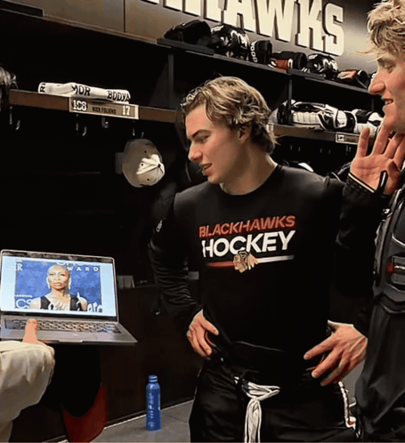 A Man Wearing A Black Hawks Hockey Shirt Stands In A Locker Room GIF