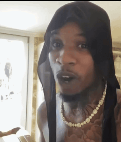 A Man Wearing A Black Head Scarf And A Pearl Necklace Looks At The Camera GIF