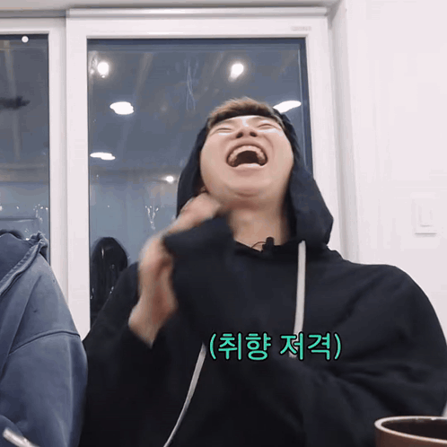 A Man Wearing A Black Hoodie Is Laughing In Front Of A Window With Korean Writing On The Bottom Right GIF