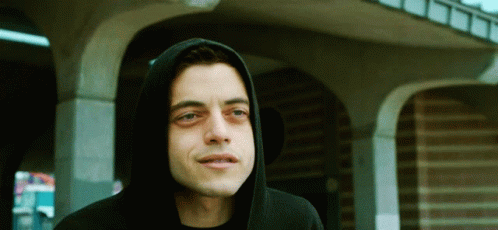 A Man Wearing A Black Hoodie Stands In Front Of A Brick Building GIF