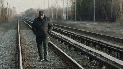 A Man Wearing A Black Jacket And A Black Hat Walks Along Train Tracks GIF